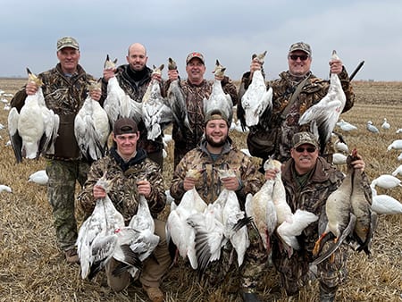 A group of hunters poses with their harvested snow geese and white-fronted geese in a field.