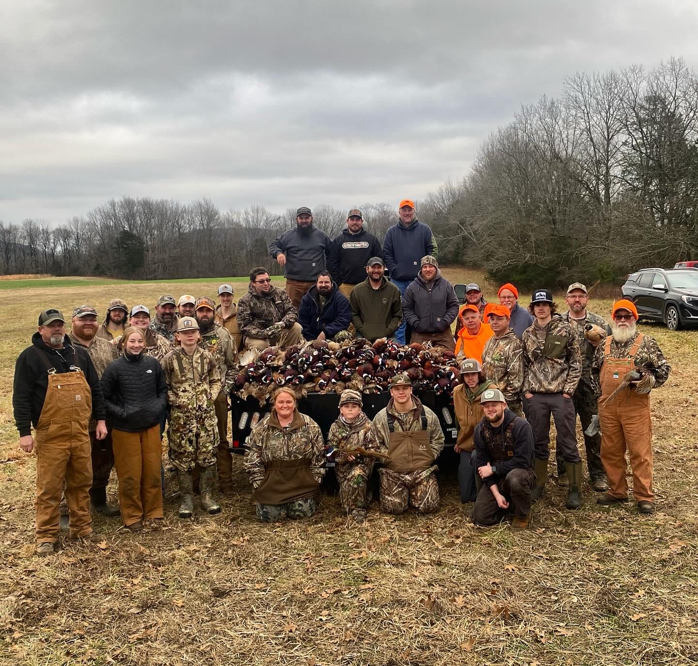 A large group of hunters poses with a pile of harvested ring-necked pheasants on a cloudy day in a field.