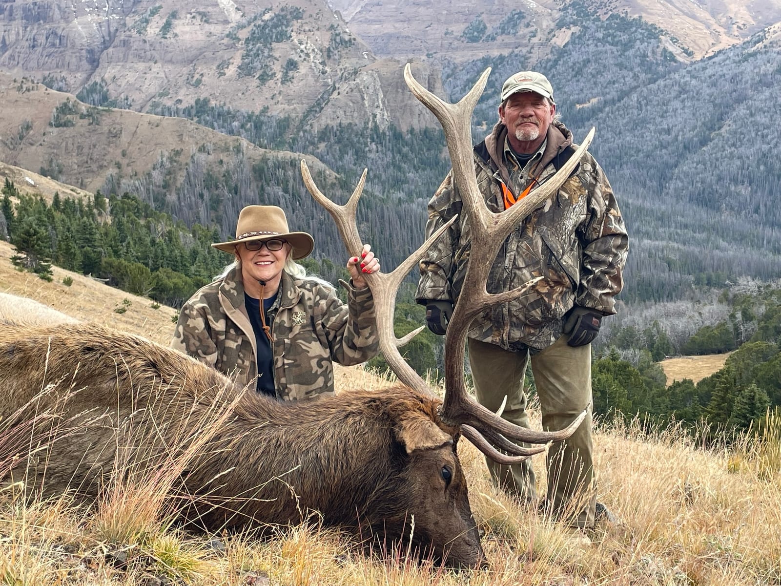 Two hunters pose with a harvested elk in a mountainous landscape.