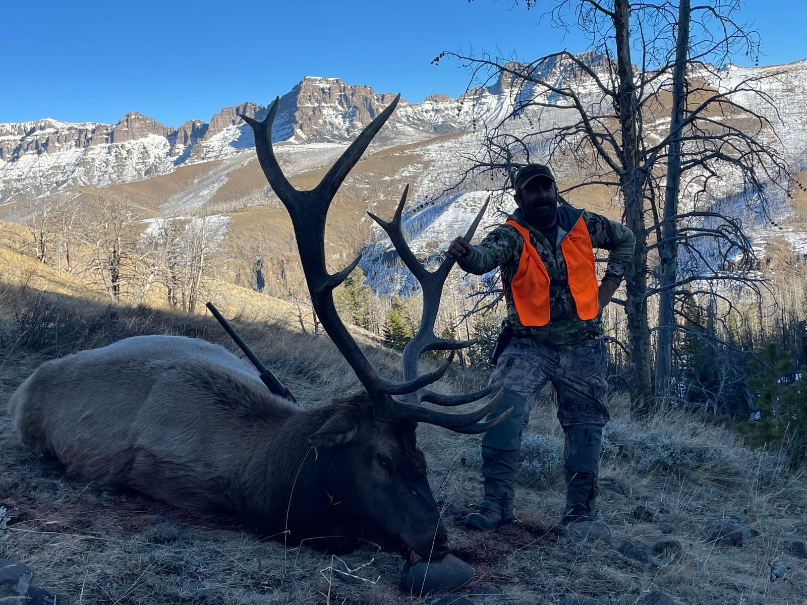 A hunter poses with a harvested elk in a mountainous, snowy landscape.