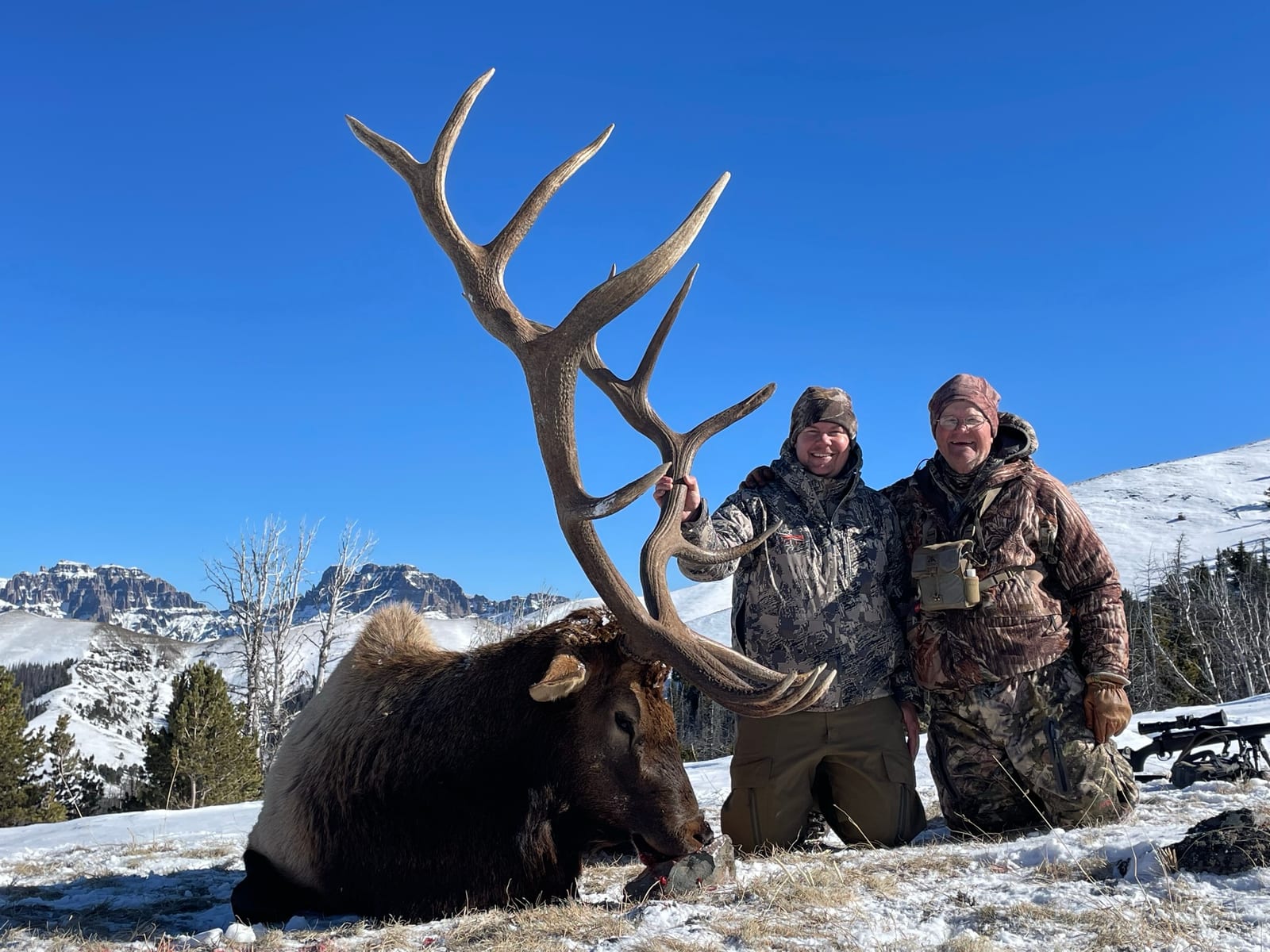 Two hunters pose with a harvested elk in a snowy, mountainous landscape.