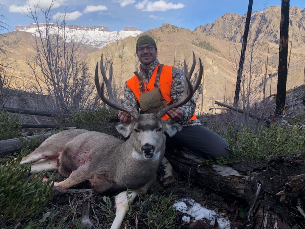 A hunter in an orange vest poses with a harvested mule deer in a mountainous, treed landscape.