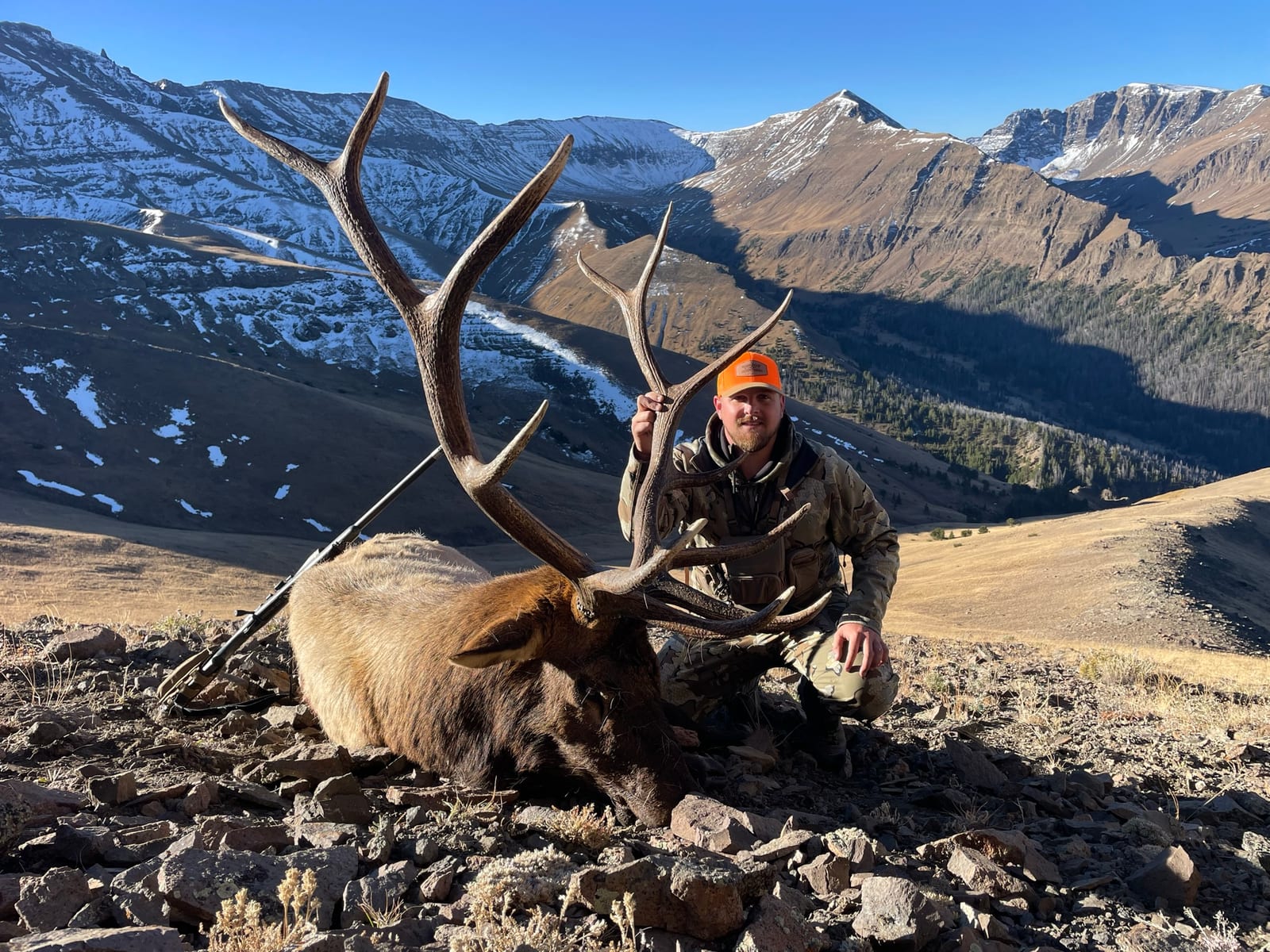 A hunter poses with a harvested elk in a mountainous, snowy landscape.