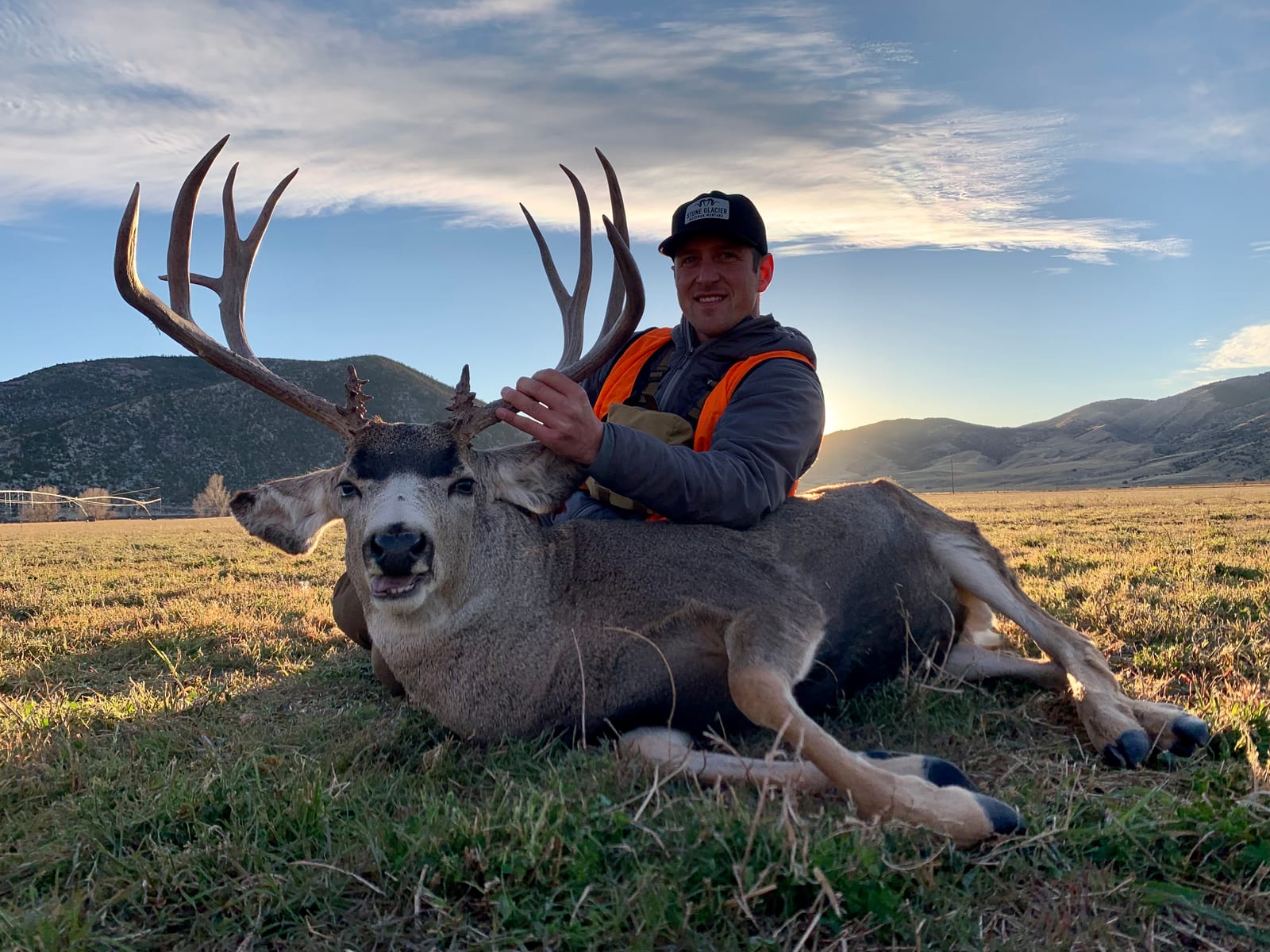 A hunter poses with a harvested mule deer in a grassy field with mountains in the background.