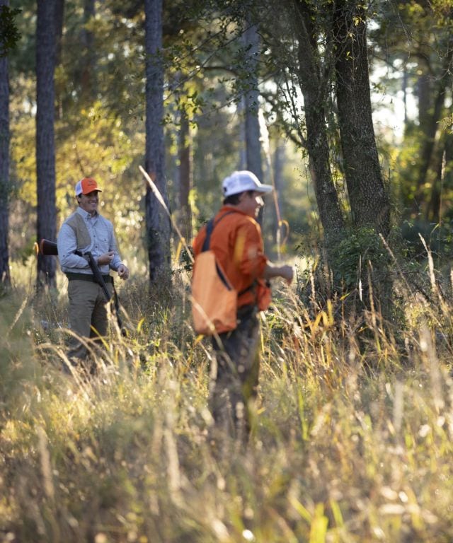 Two hunters walk through tall grass and trees in a sunny field.