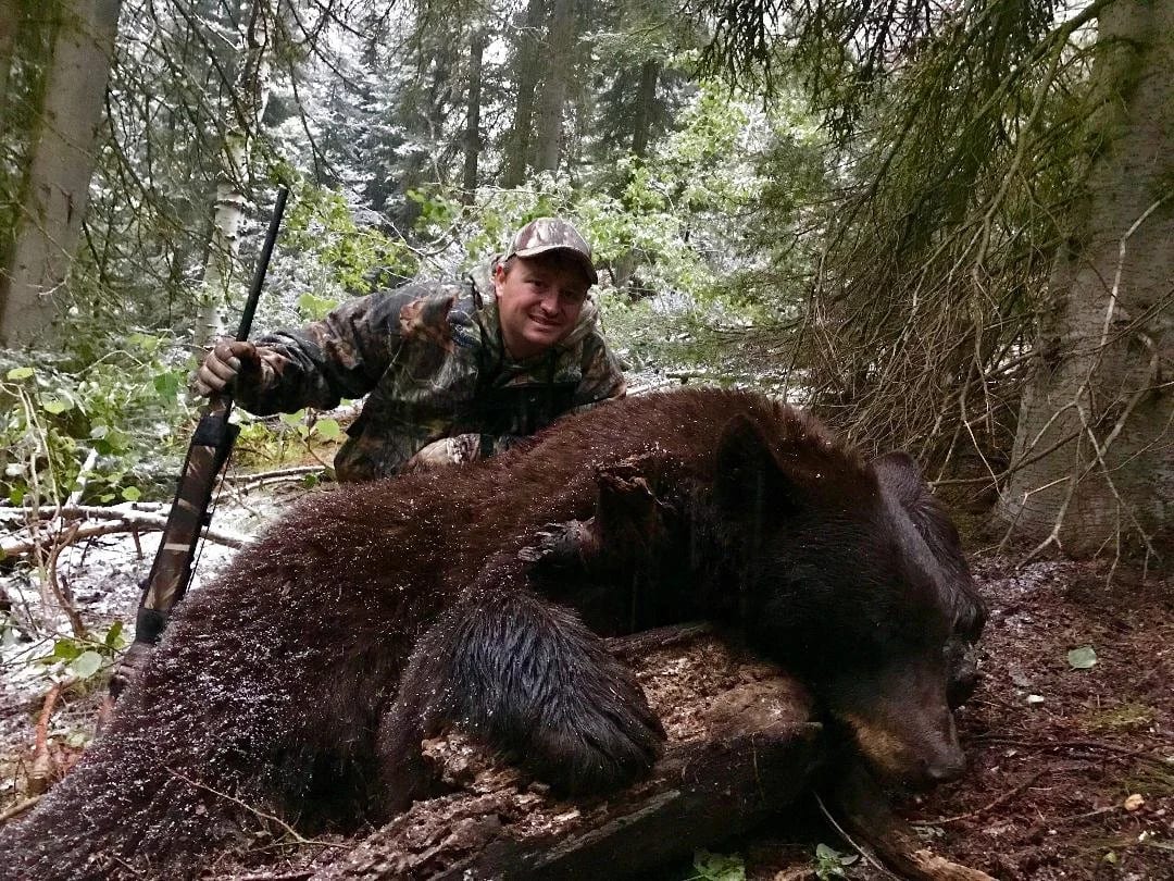 A hunter in camouflage kneels in a snowy forest, holding a rifle beside a large black bear lying on a fallen log.