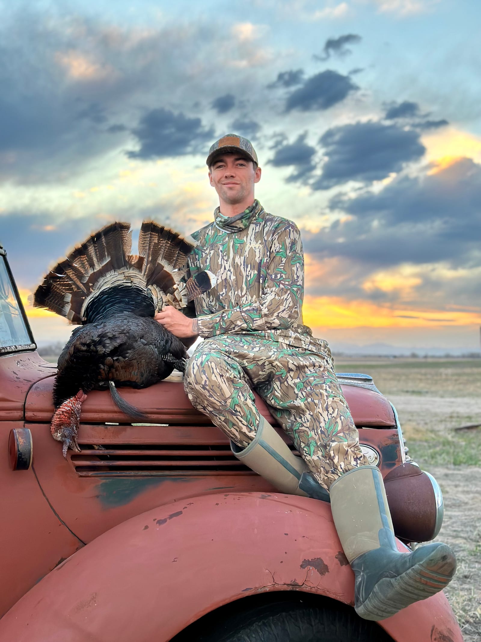 A hunter in camouflage sits on the hood of an old truck, holding a harvested wild turkey at sunset.