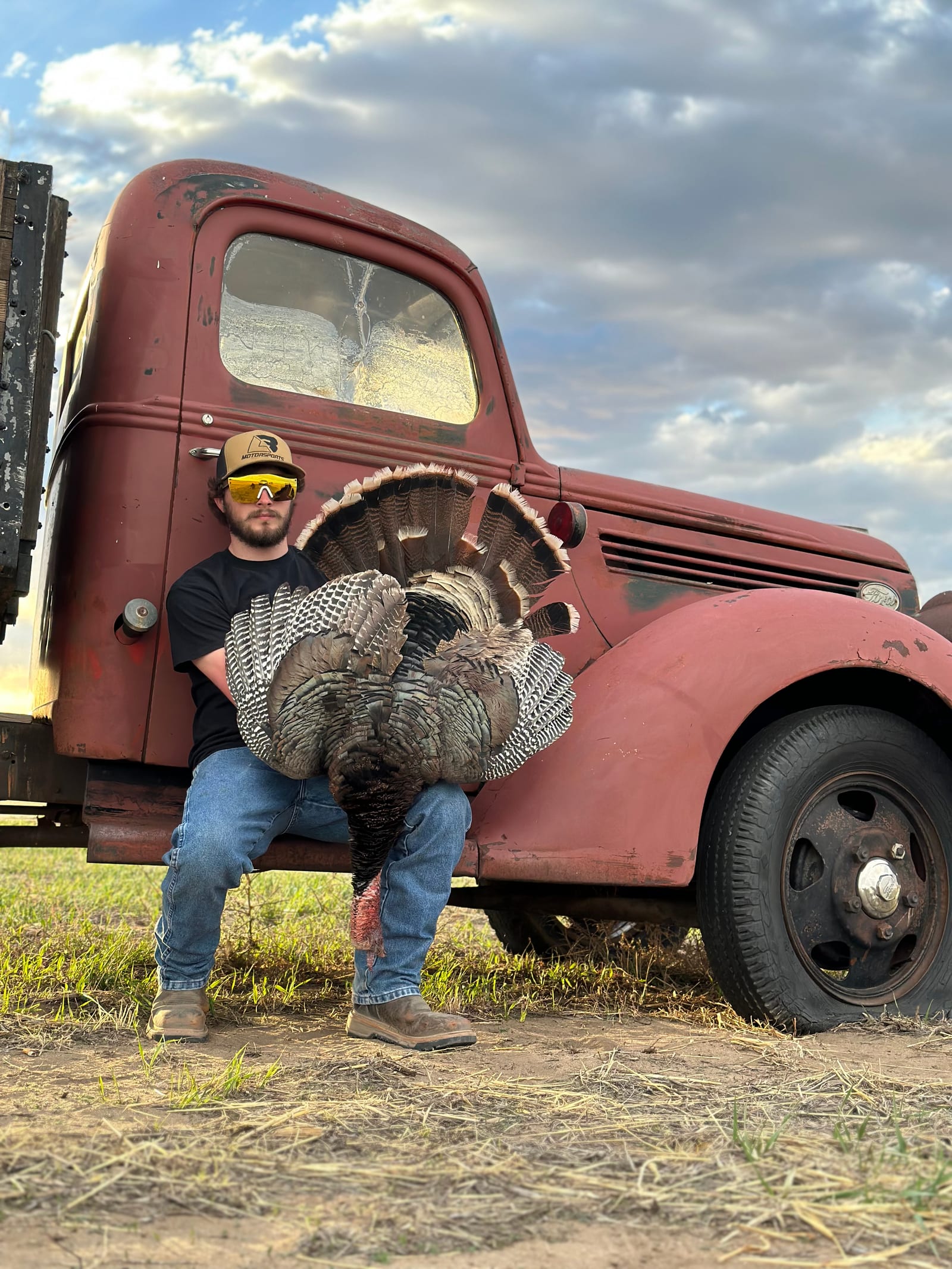 A hunter sits on the running board of an old red truck, holding a harvested wild turkey.
