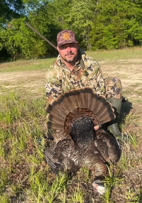 A hunting enthusiast displaying a wild turkey with a shotgun in an outdoor setting.