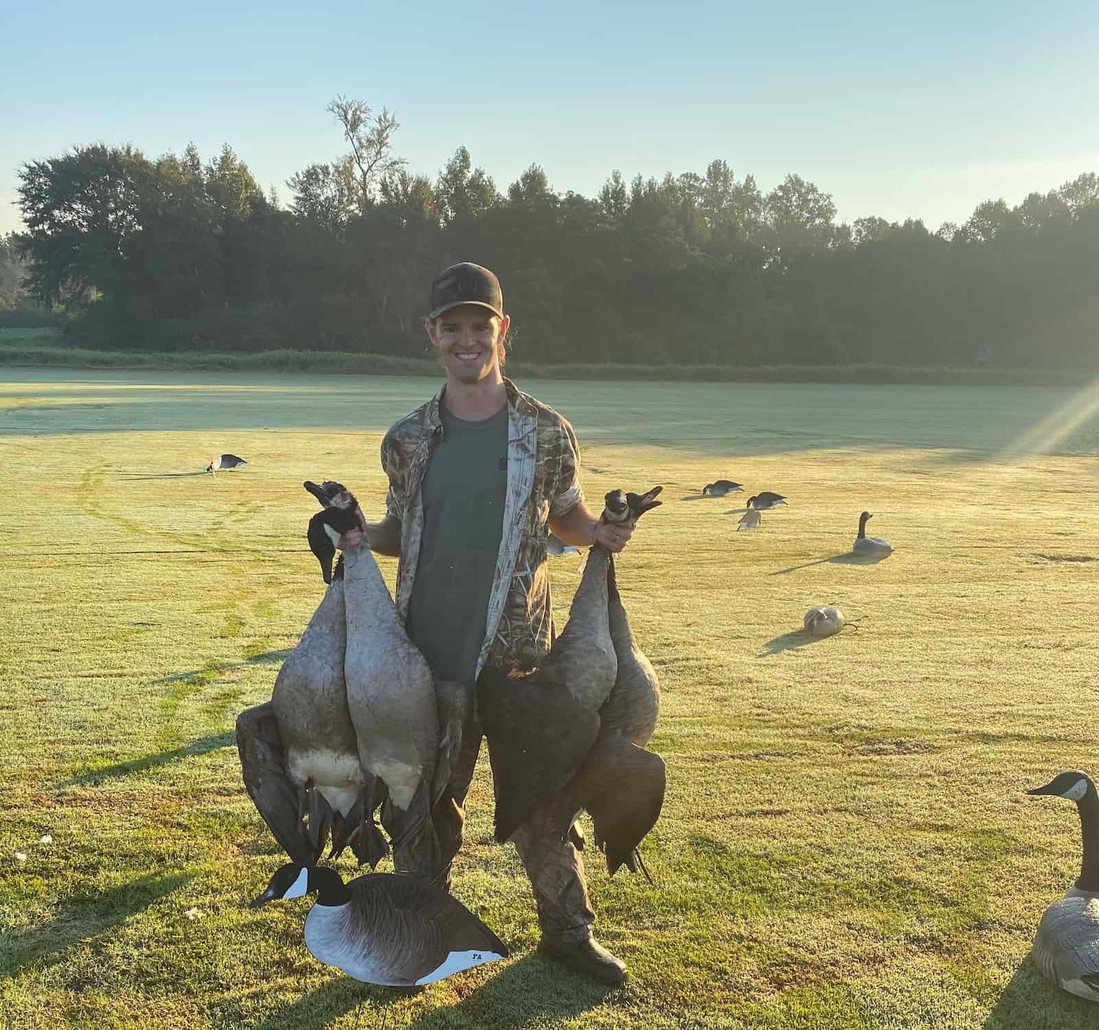 A hunter smiles while holding several harvested geese in a grassy field with decoys and trees in the background.