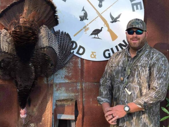 A hunter in camouflage stands next to a harvested wild turkey.