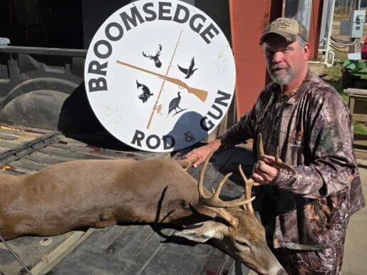 Man with deer and Broomsedge Rod & Gun sign outside store.