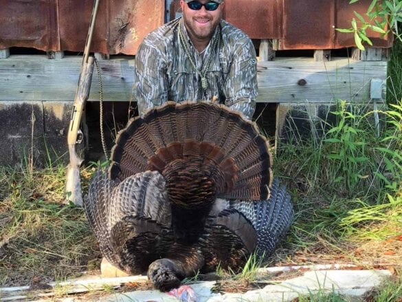 A man in camouflage poses with a harvested wild turkey and a shotgun.