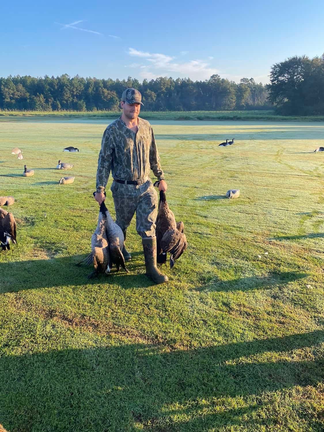 A man in camouflage holds two harvested geese in a grassy field with decoys.