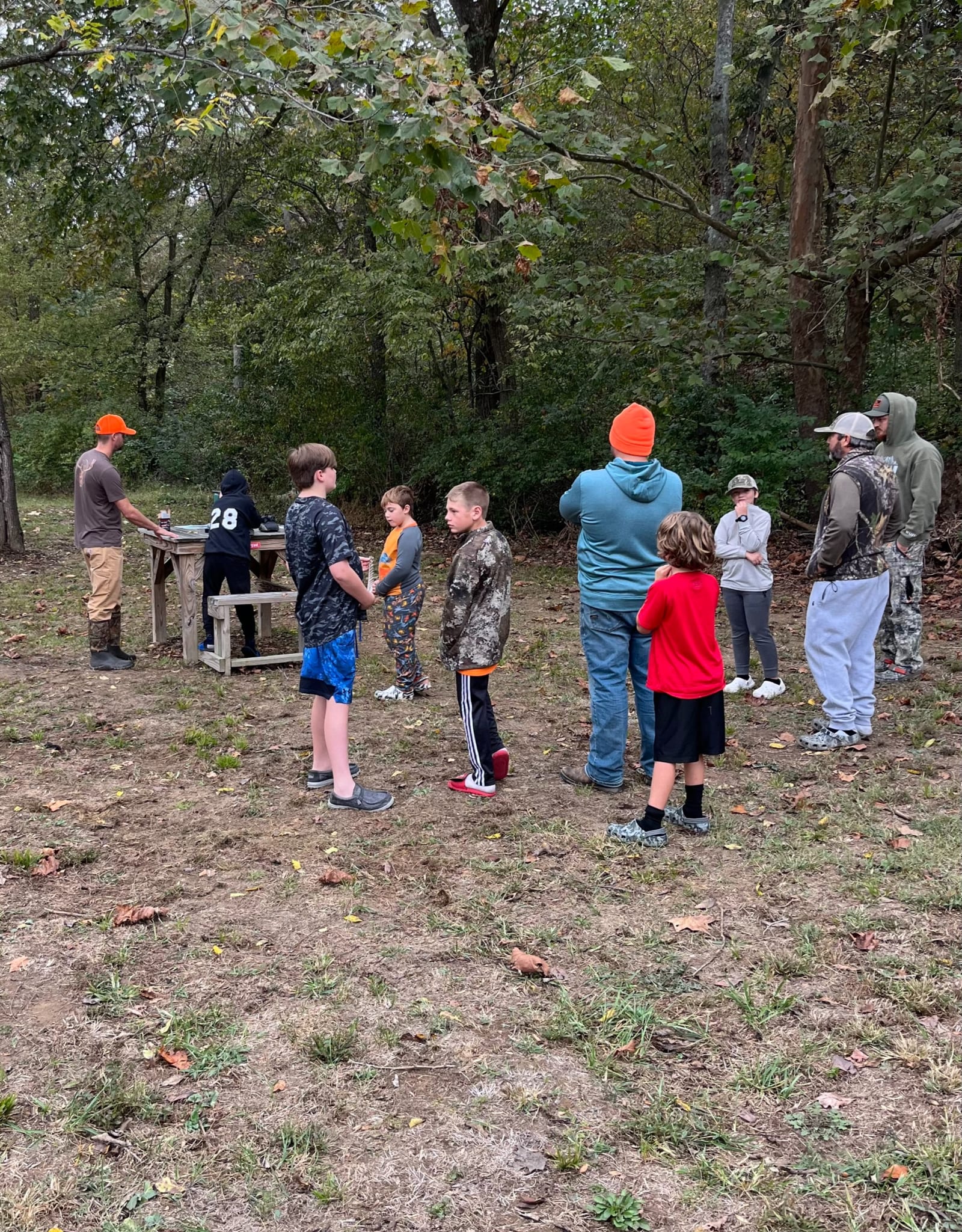 A group of people, including adults and children, are gathered outdoors in a wooded area.