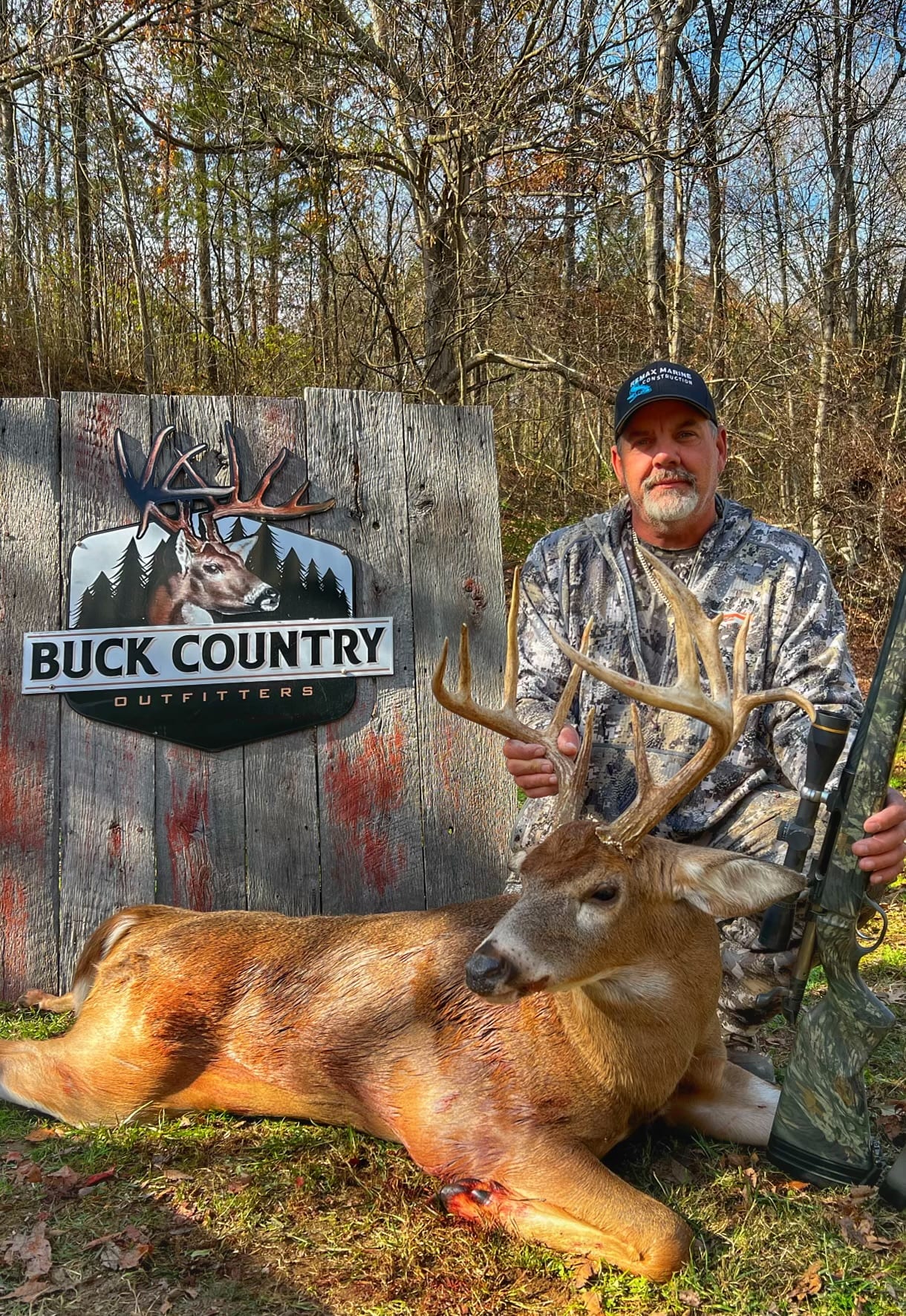 A hunter poses with a harvested whitetail deer in front of a "Buck Country Outfitters" sign.