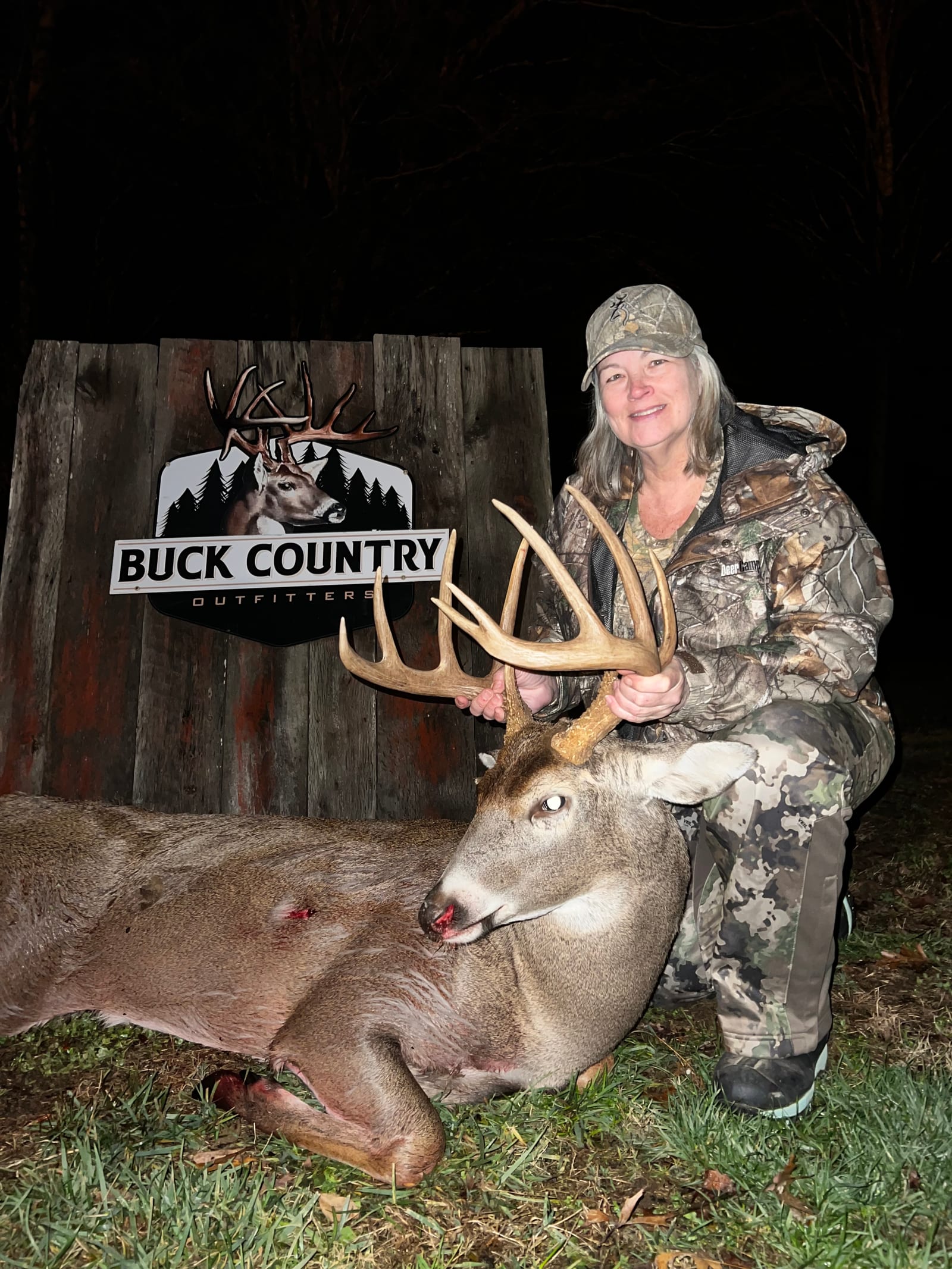 A woman in camouflage poses with a harvested whitetail deer at night next to a Buck Country Outfitters sign.