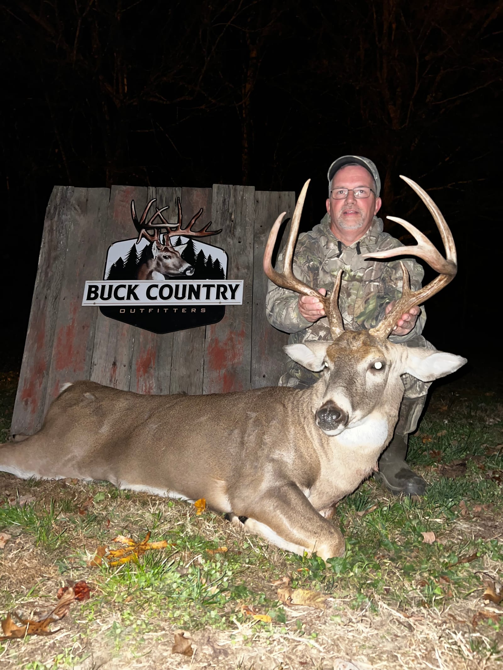 A hunter poses with a harvested whitetail deer at night in front of a "Buck Country Outfitters" sign.