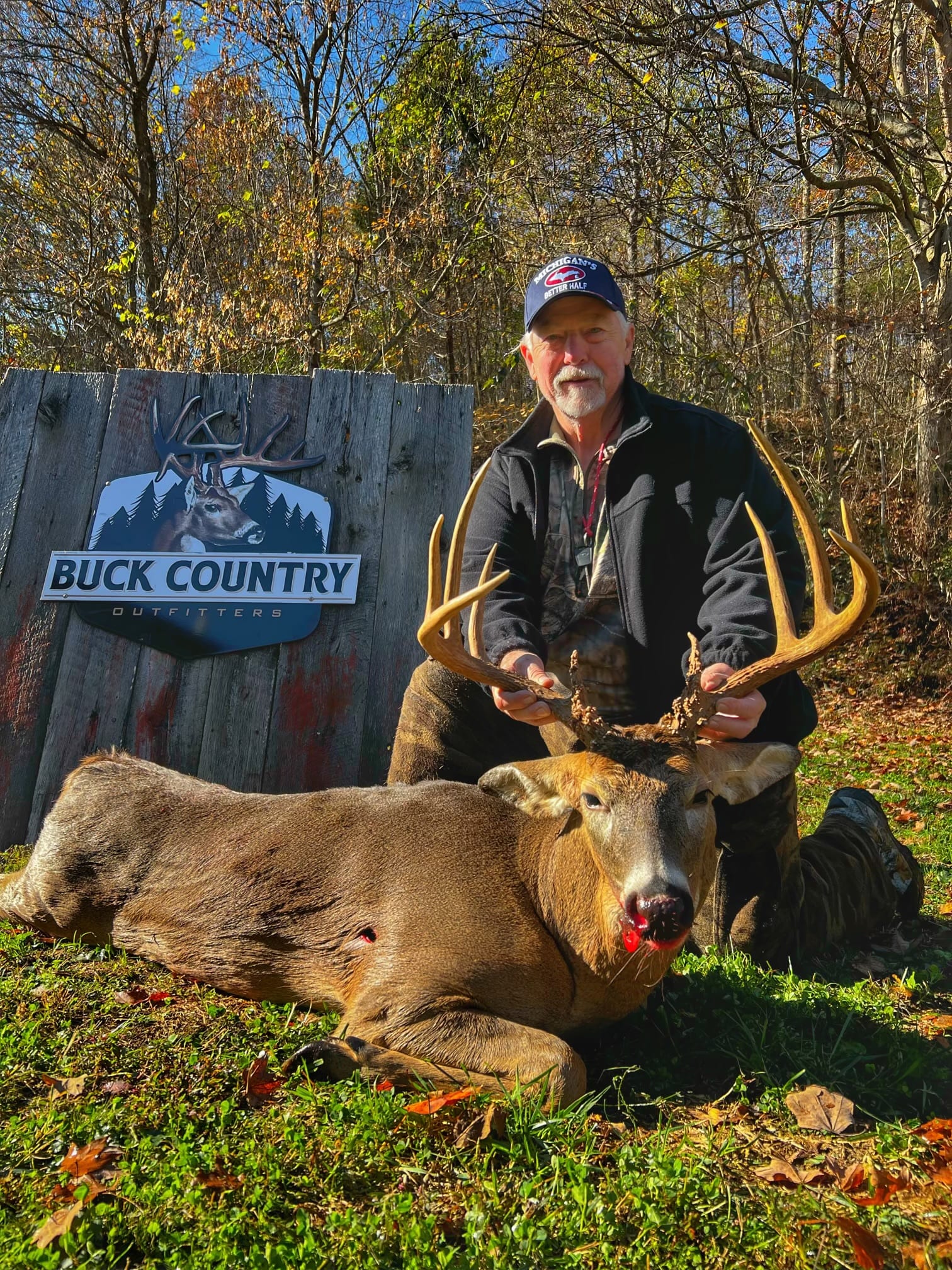 A hunter kneels next to a harvested whitetail deer in front of a "Buck Country Outfitters" sign.