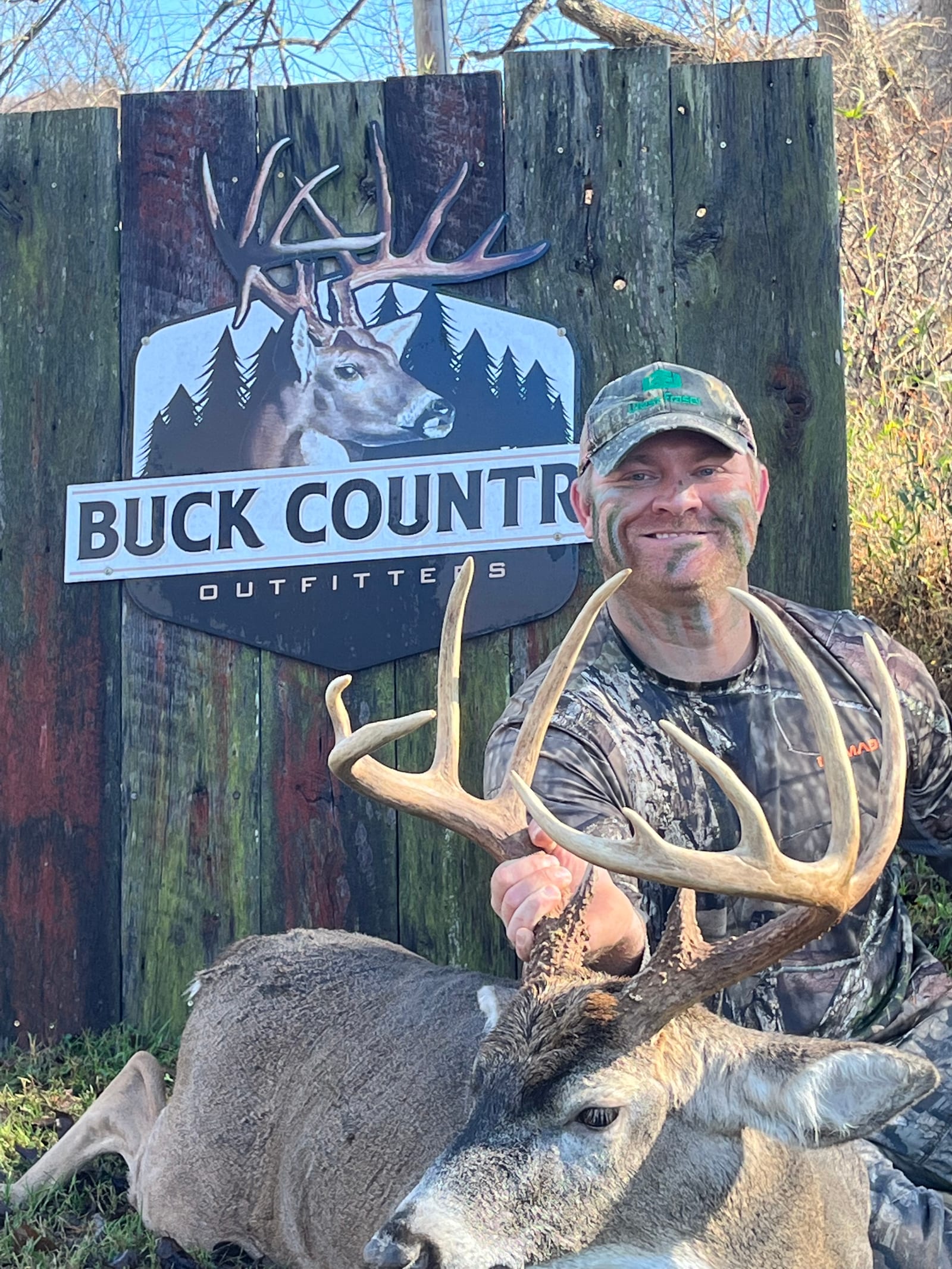 A man in camouflage with face paint poses with a harvested whitetail deer in front of a "Buck Country Outfitters" sign.