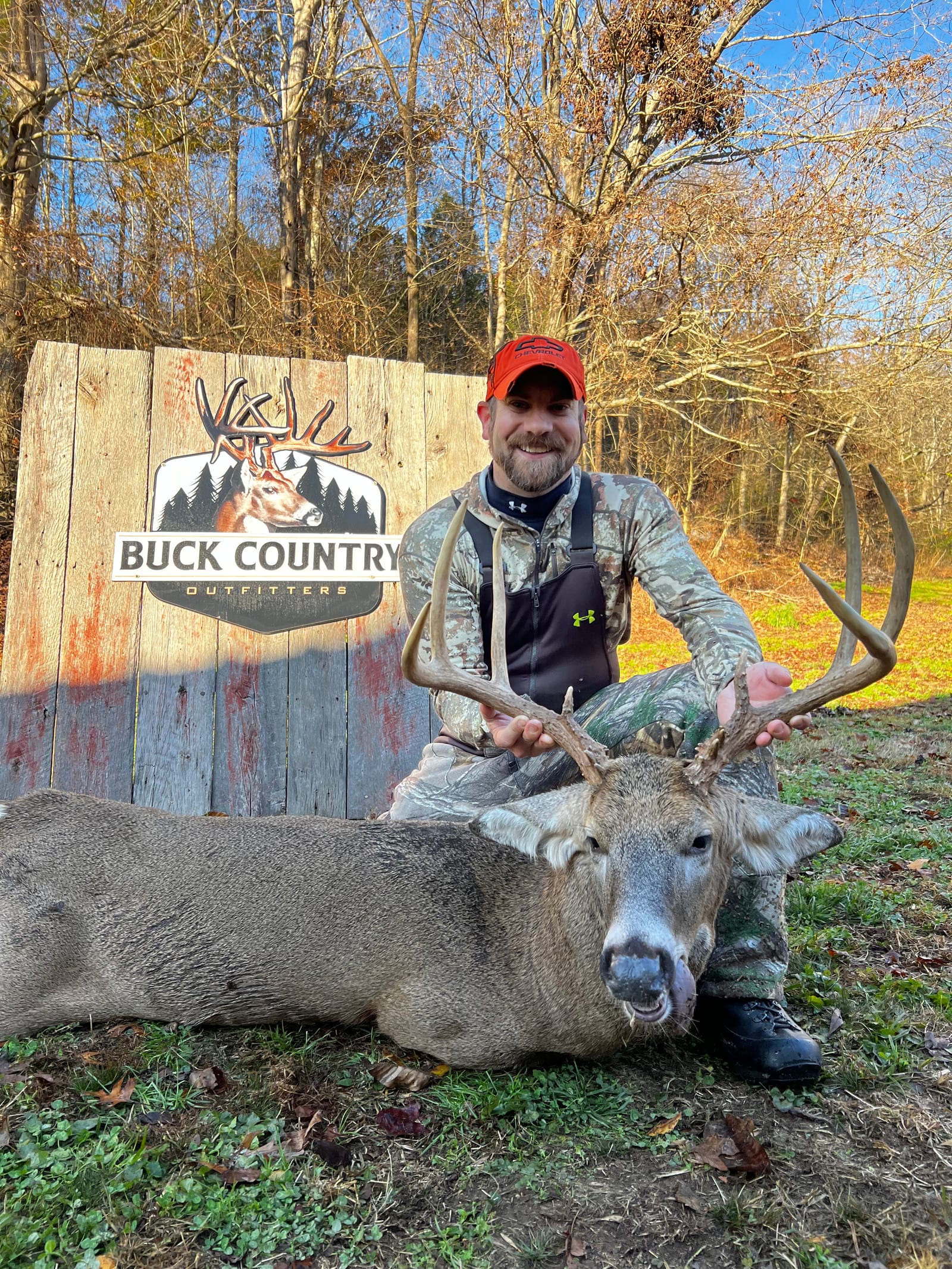 A hunter poses with a harvested whitetail deer in front of a "Buck Country Outfitters" sign.