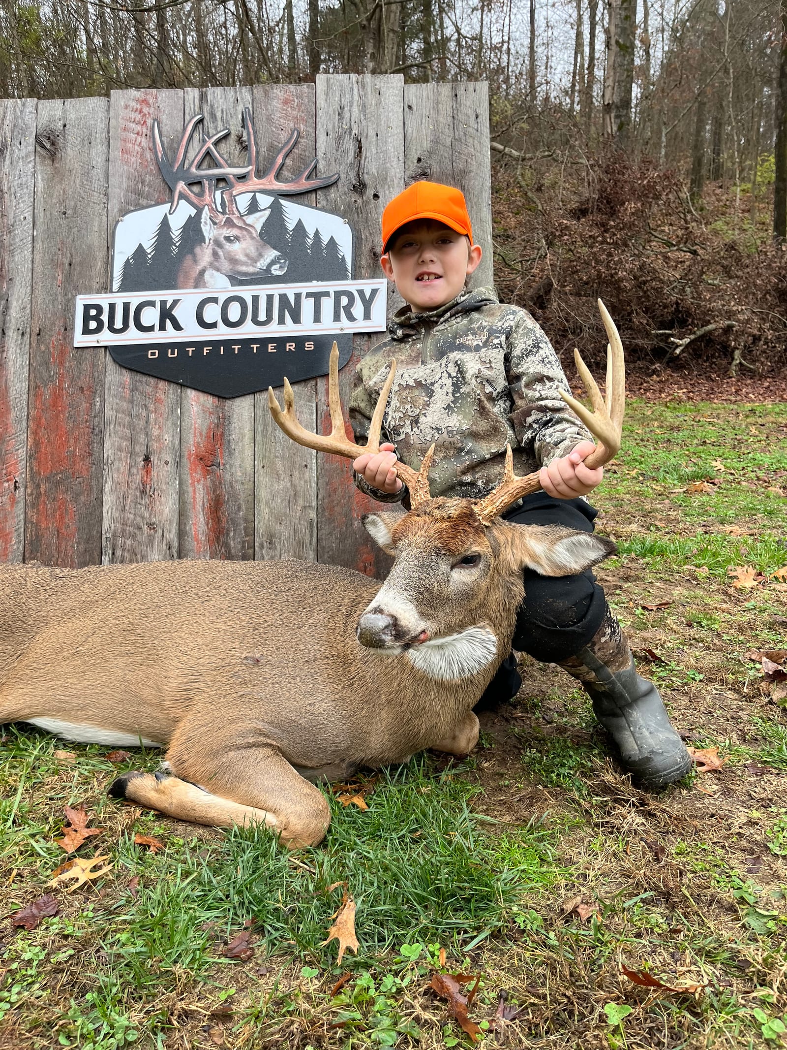 A young boy poses with a harvested whitetail deer in front of a 'Buck Country Outfitters' sign.