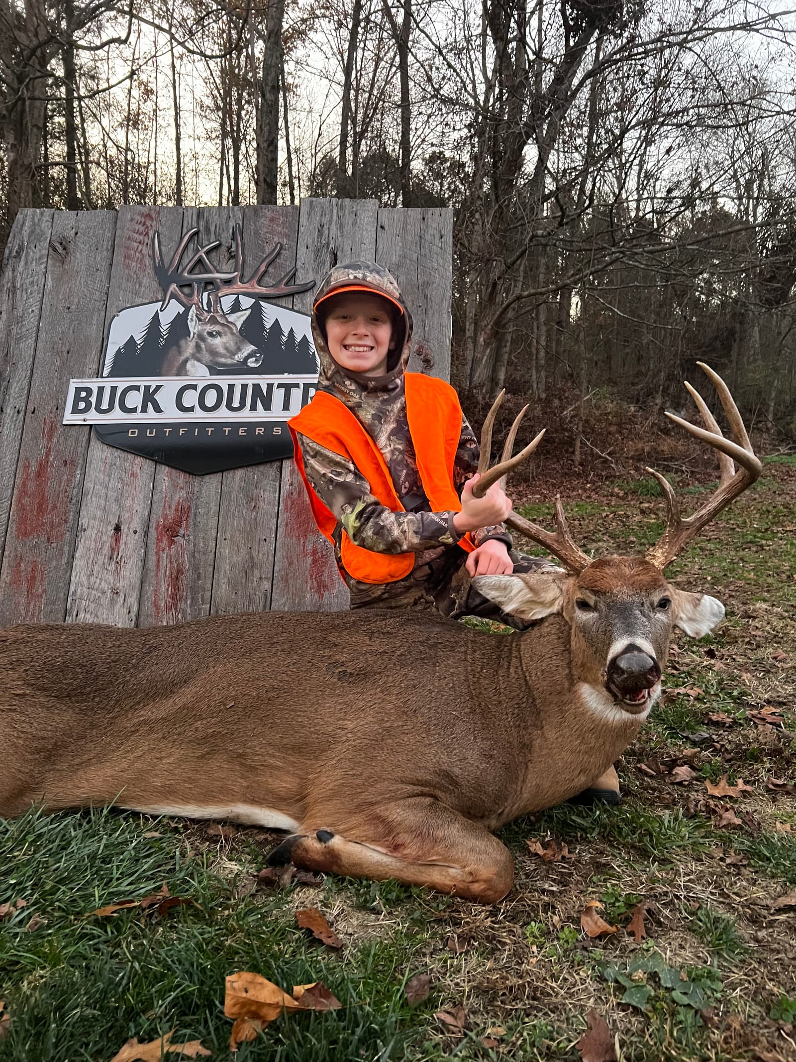 A young boy in hunting attire poses with a harvested whitetail deer in front of a "Buck Country Outfitters" sign.