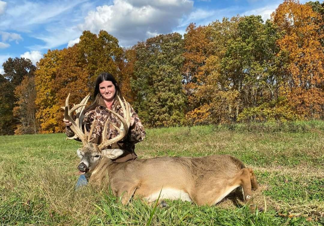 A woman poses with a harvested whitetail deer in a grassy field with autumn trees in the background.