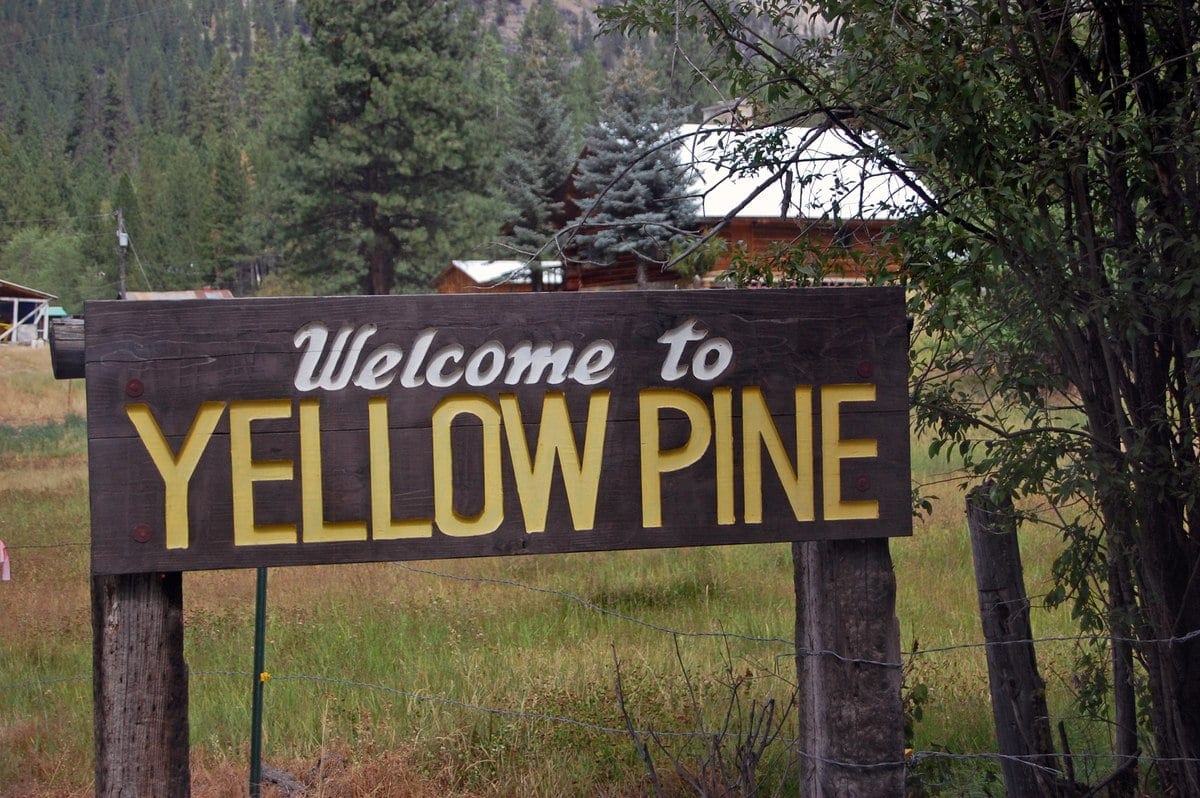 A wooden sign reads 'Welcome to YELLOW PINE' in a natural setting with trees and buildings in the background.