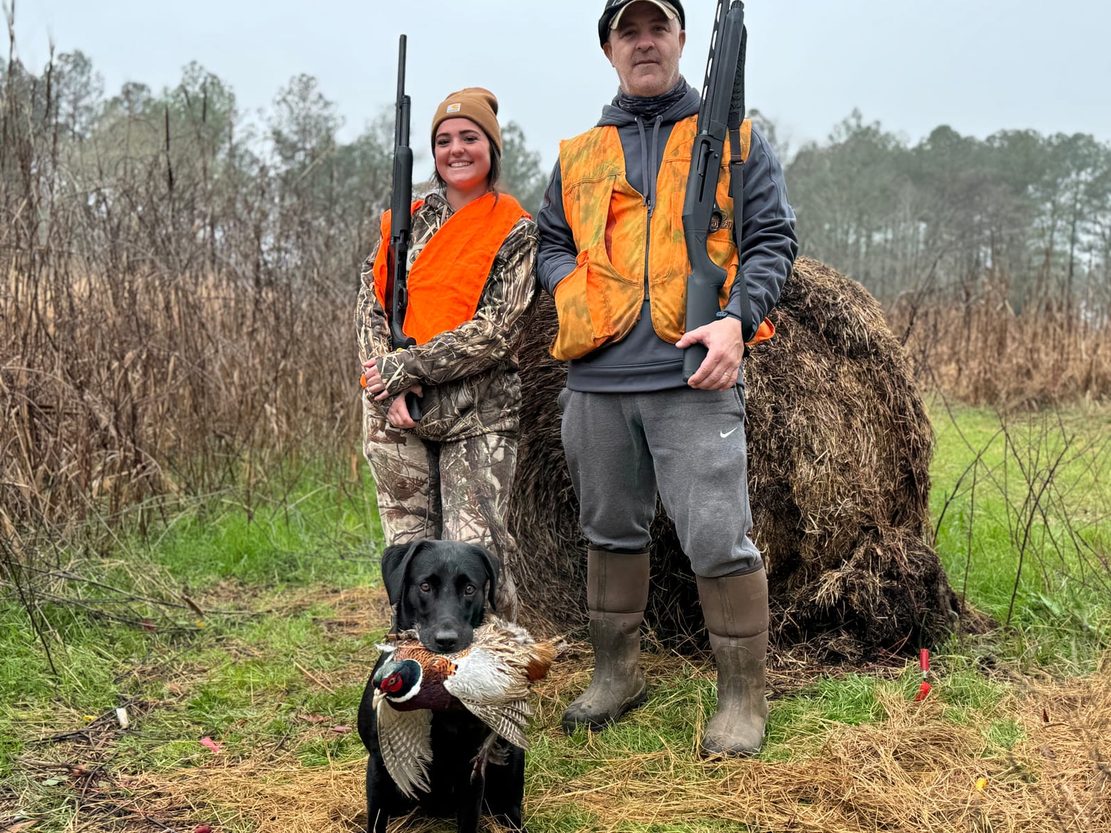 Two hunters and a black dog pose with a harvested ring-necked pheasant in a field.