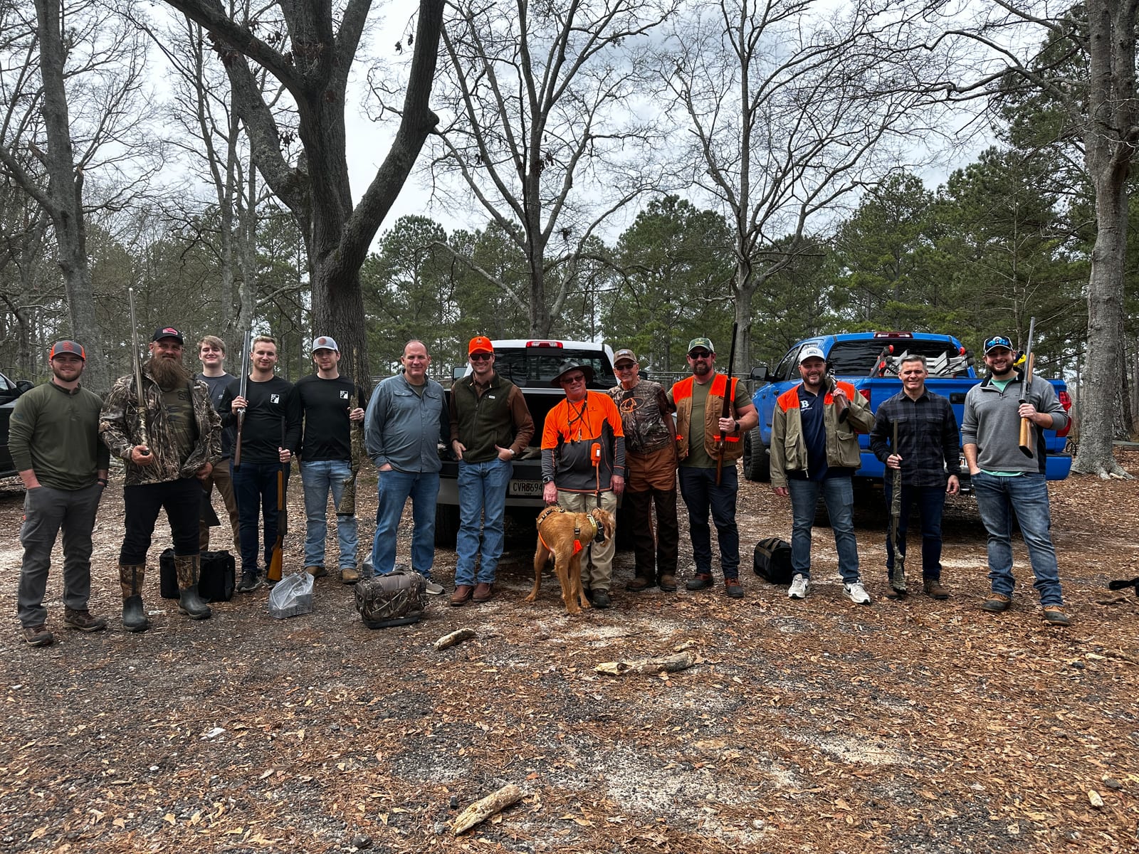 A group of hunters and a dog pose with a harvested bird in a wooded area with trucks.