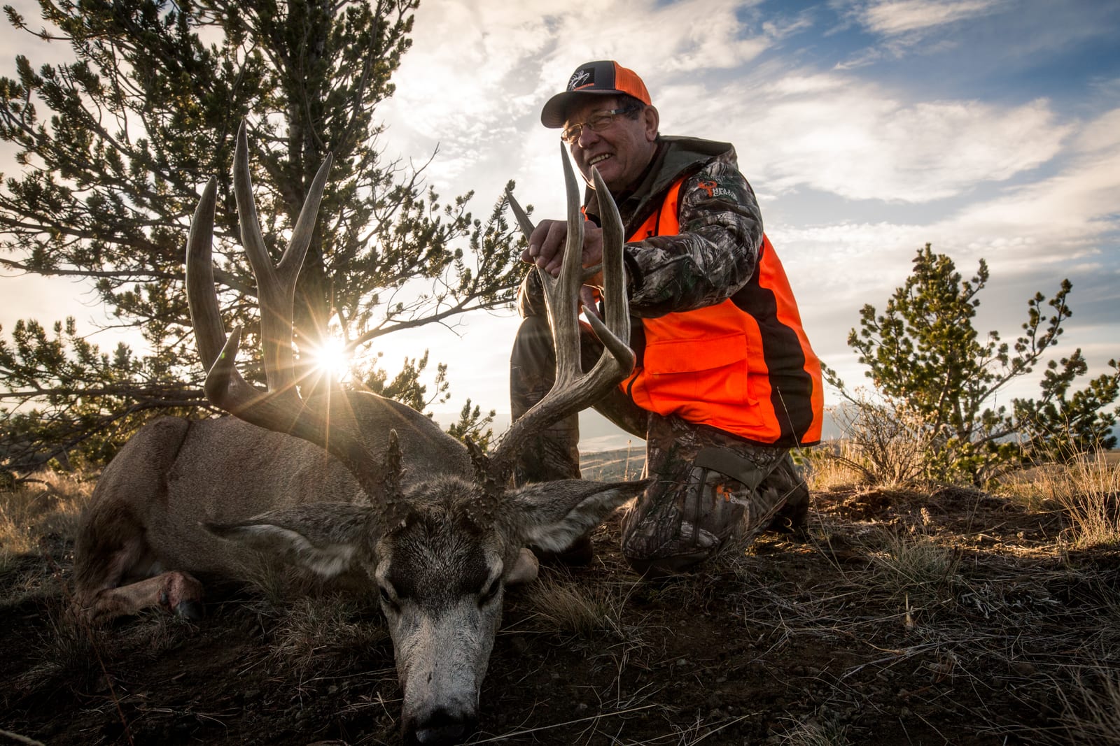 A hunter kneels beside a harvested mule deer with large antlers, with the sun setting behind them.