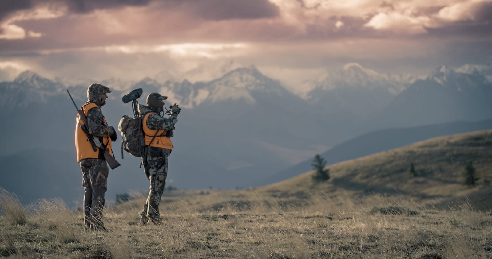 Two hunters stand in a grassy field, one with a rifle and the other with binoculars, looking out at a vast landscape of snow-capped mountains under a dramatic sky.