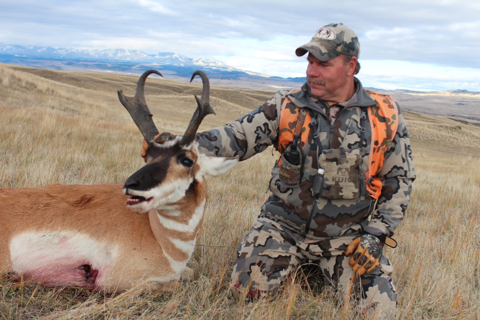A hunter kneels next to a harvested pronghorn in a grassy field with mountains in the background.