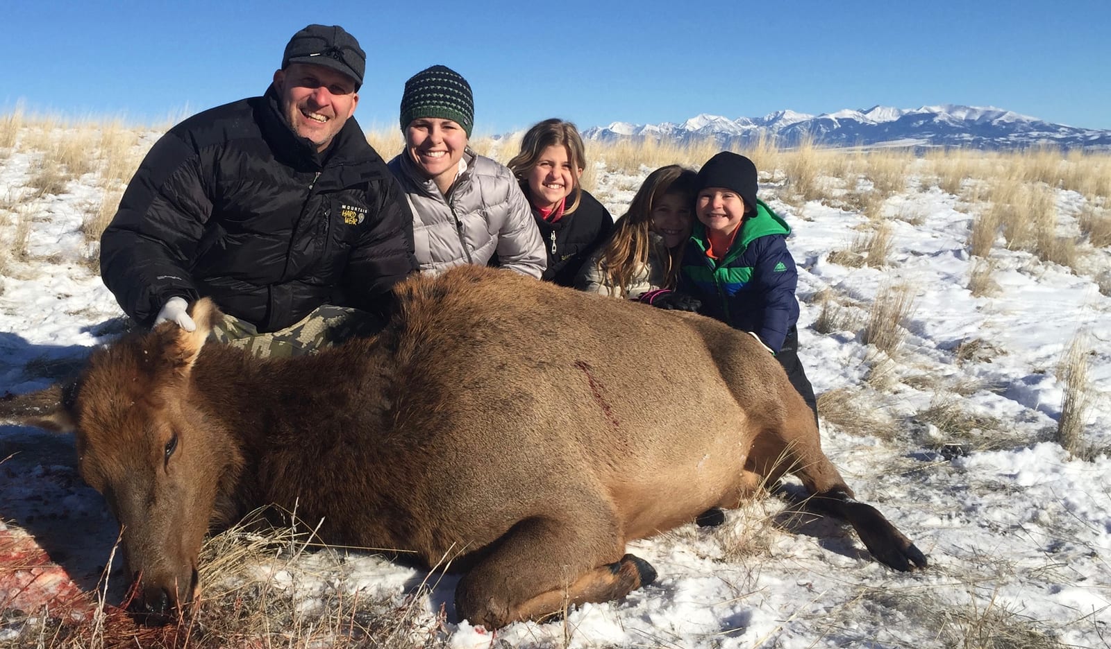 A group of people pose with a harvested elk in a snowy field with mountains in the background.