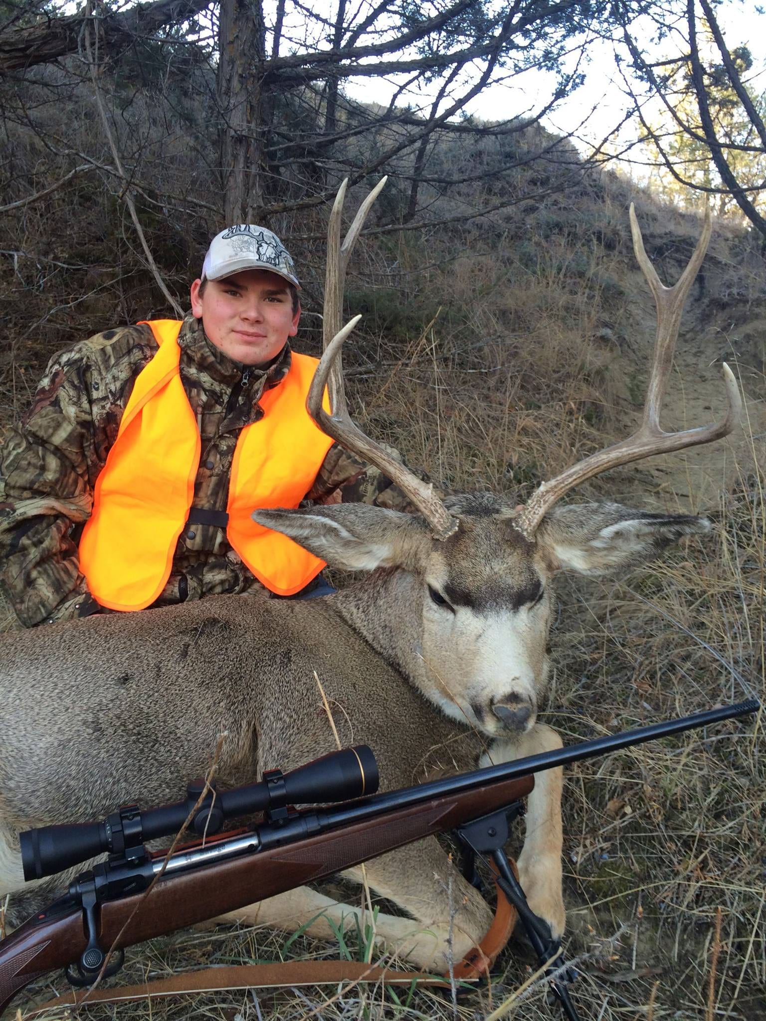 A hunter in camouflage and an orange vest poses with a harvested mule deer and a rifle.