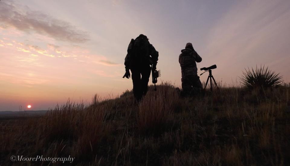 Two hunters or guides are silhouetted against a sunset or sunrise sky on a grassy hill, one standing and one kneeling with a spotting scope.