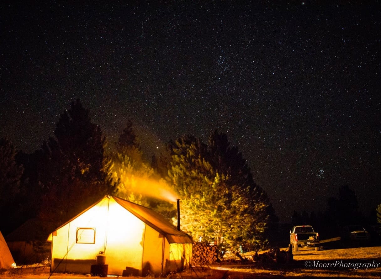 A brightly lit tent camp under a starry night sky, with trees and a truck in the background.