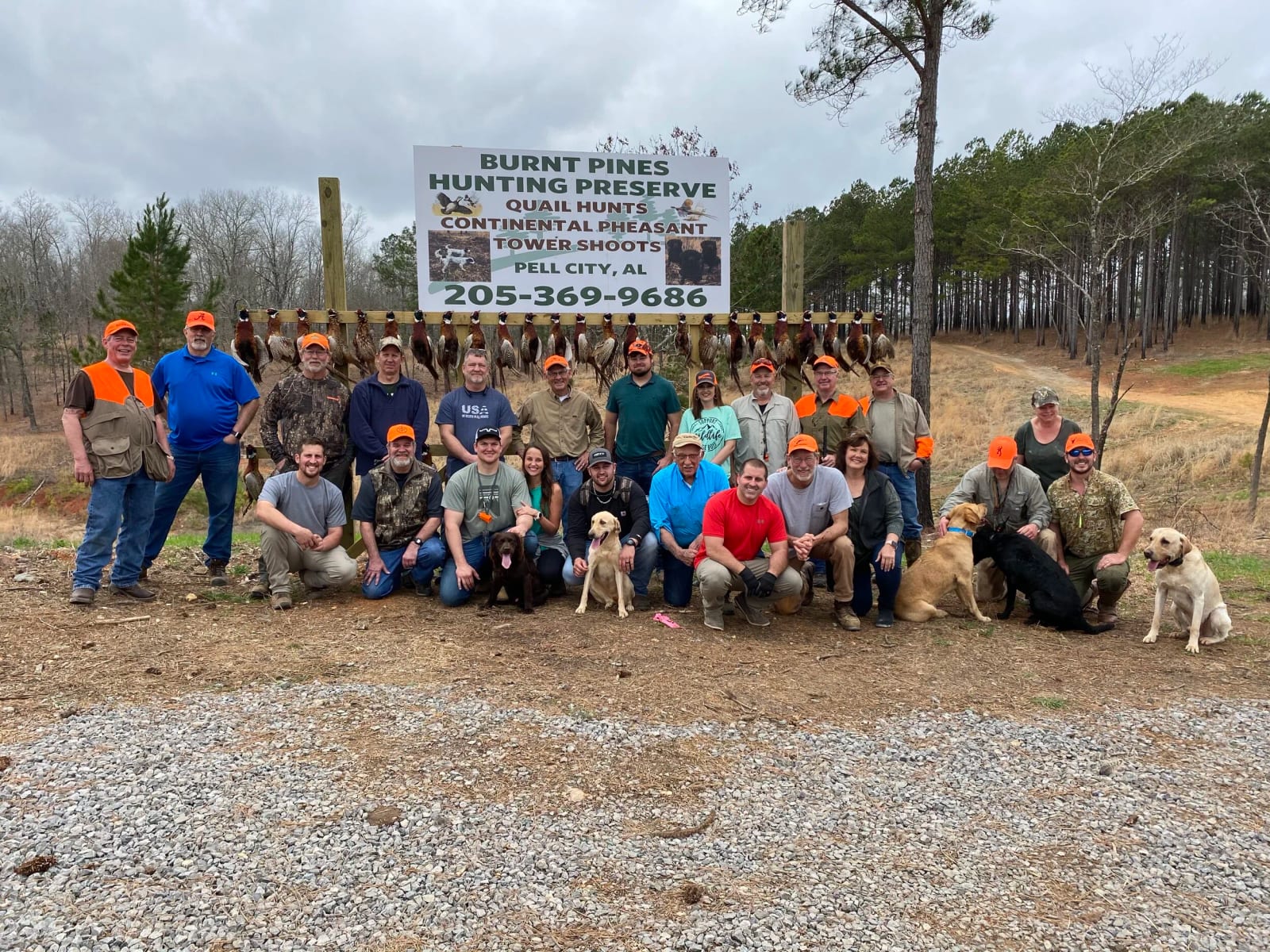 Group of hunters and dogs posing in front of a sign at Burnt Pines Hunting Preserve, with quail hanging on a line behind them, in Pell City, Alabama.