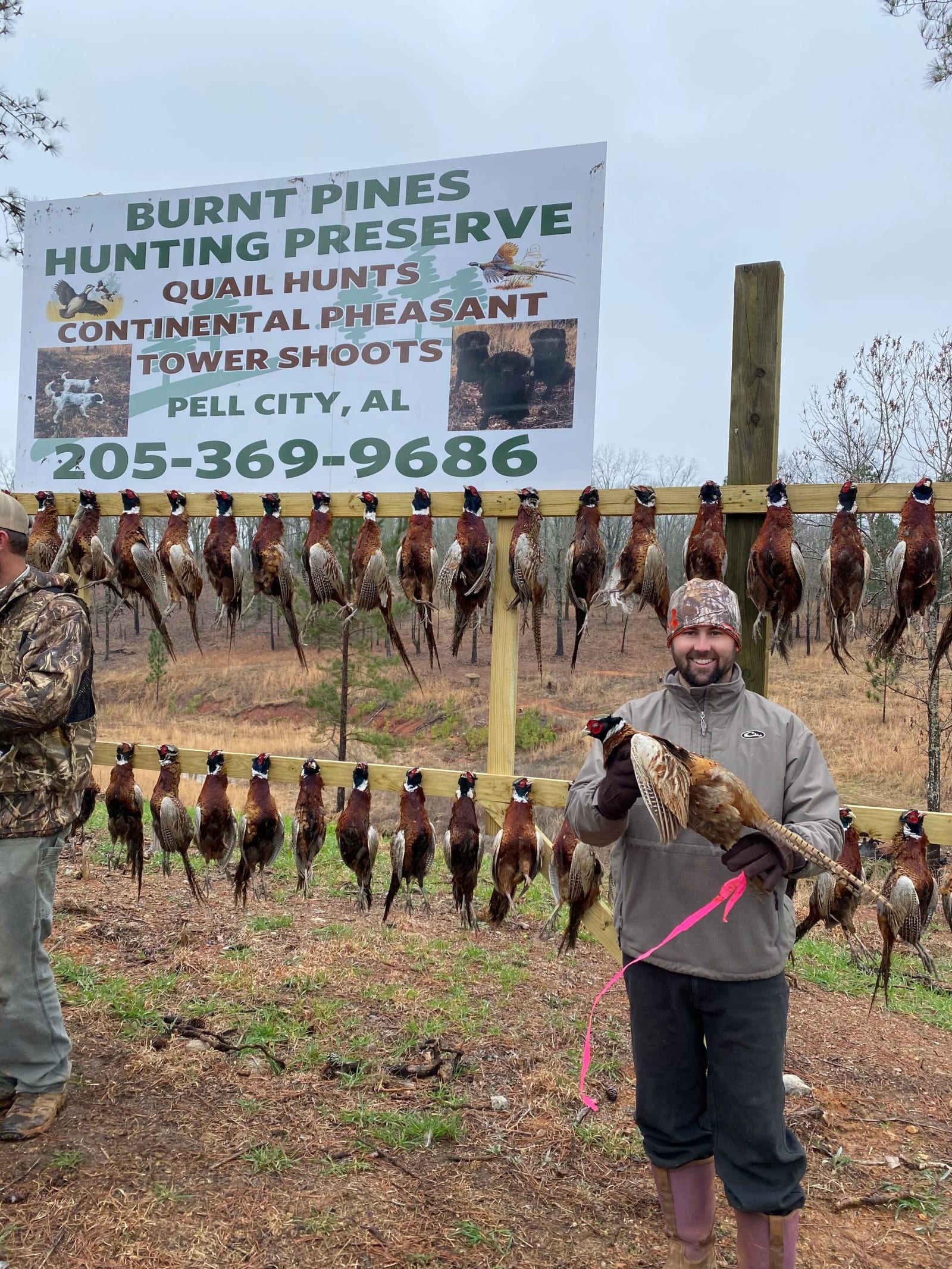 Man holding a pheasant in front of a display of hanging pheasants at Burnt Pines Hunting Preserve in Pell City, Alabama.