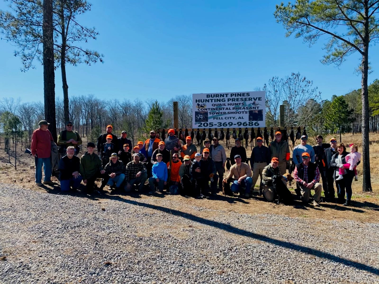 Group of people outdoors in a wooded area, some wearing orange hats, with a sign for Burnt Pines Hunting Preserve in the background.