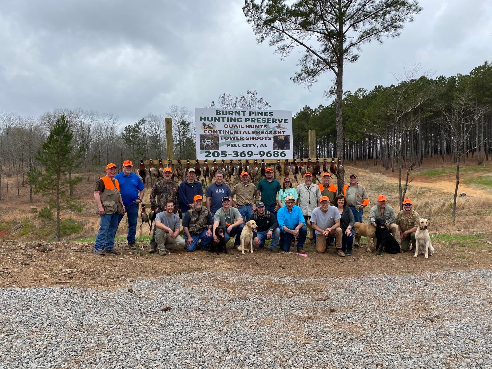 Group of people with hunting dogs and gear standing in front of a sign at Burnt Pines Hunting Preserve in Pell City, Alabama, with trees and a cloudy sky in the background.
