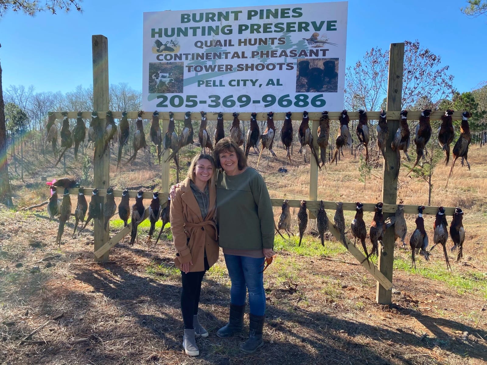 Two women standing together outdoors in front of a sign and a display of hanging quail birds. The sign advertises Burnt Pines Hunting Preserve for quail hunts and other outdoor activities in Pell City, Alabama.
