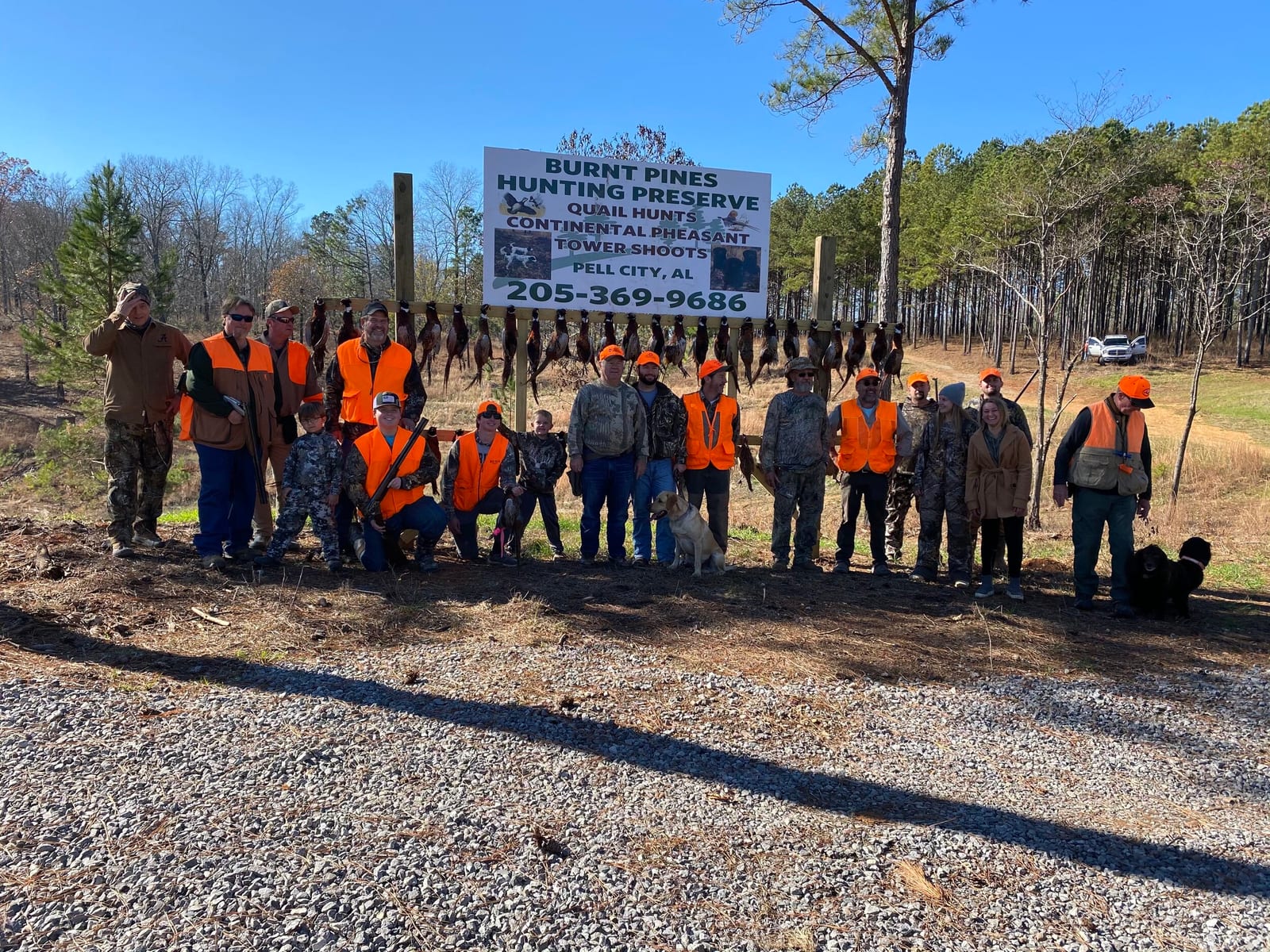 Group of people in hunting gear standing in front of a sign that reads "Burnt Pines Hunting Preserve" with ducks hanging on a line above them. Some individuals wear orange safety vests, and two dogs are present. The background shows a wooded area and