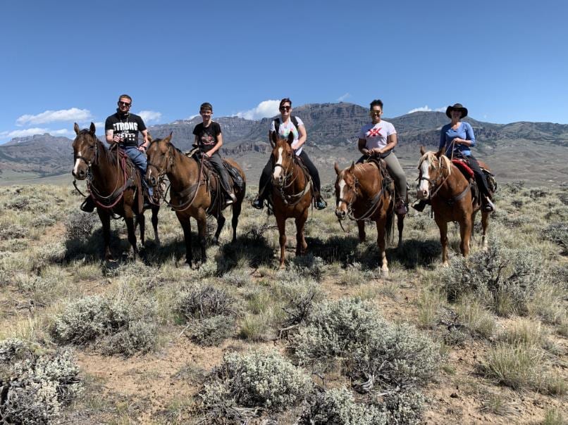 Five people are riding horses on a trail in a dry, brushy landscape with mountains in the background under a clear blue sky.