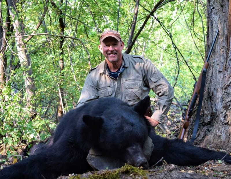 A hunter poses with a harvested black bear in a forest.