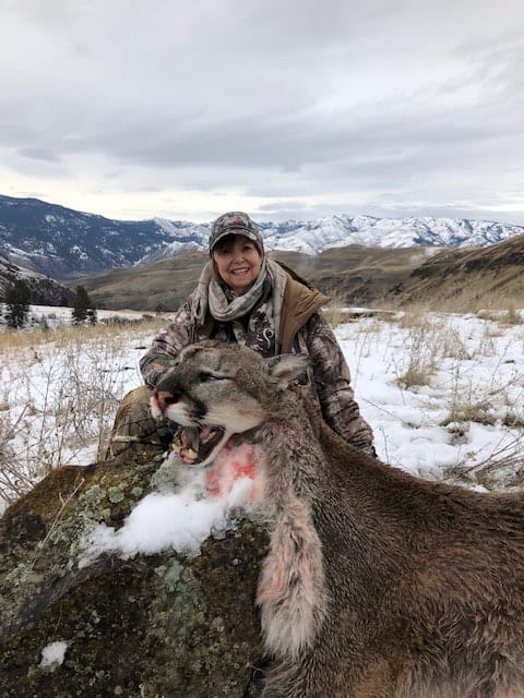 A hunter poses with a harvested mountain lion in a snowy, mountainous landscape.
