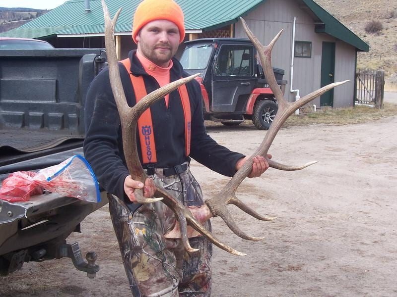 A man in hunting attire holds a pair of elk antlers in front of a building and a vehicle.