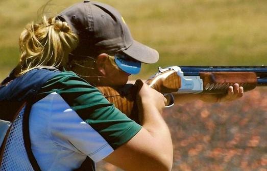 A person wearing a cap and safety glasses aims a shotgun.