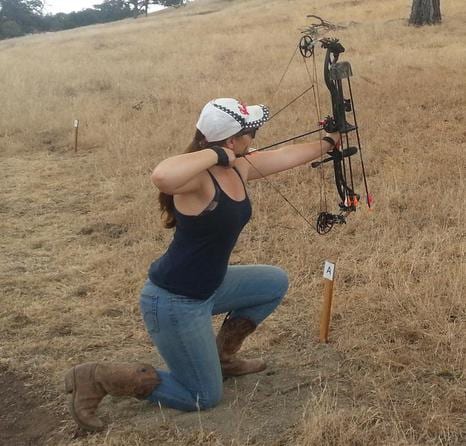 A woman kneels in a dry field, aiming a compound bow.